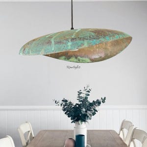 Könnte beinhalten: Eine große, ovale Pendelleuchte mit Kupferoberfläche und türkisfarbener Patina. Das Licht hängt an einem schwarzen Kabel über einem Esstisch. Die Leuchte ist ein einzigartiges, künstlerisches Statement.