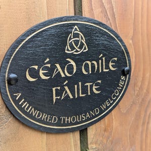 May include: Oval black slate welcome sign with gold lettering and a Celtic knot design. The sign reads "Céad Míle Fáilte" and "A Hundred Thousand Welcomes". Two black screws secure the sign to a wooden surface.