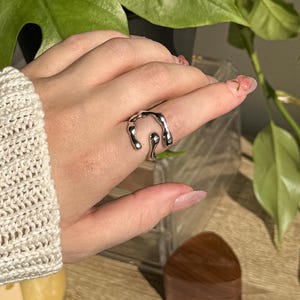 Op de afbeelding: Een zilverkleurige ring met een uniek, vloeiend ontwerp wordt aan een vinger gedragen. De ring heeft een abstracte vorm met gebogen lijnen en een gepolijste afwerking. De achtergrond omvat een groene plant, een houten doos en een kleine kaart.