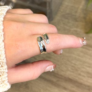 May include: A silver ring with an unusual, open design. The ring wraps around the finger with a smooth, reflective surface. The jewellery is shown on a hand with a cream-coloured sweater in the background.