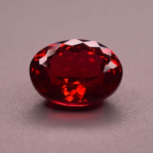 Könnte beinhalten: Ein tiefroter, oval geformter Edelstein mit facettierter Oberfläche. Der Stein weist einen Brillantschliff auf, der das Licht auf seiner polierten, durchscheinenden Oberfläche reflektiert und eine satte, kräftige karmesinrote Farbe zeigt.