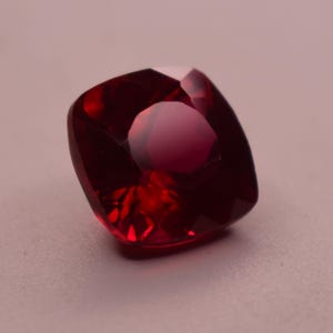 Puede incluir: Una piedra preciosa de color rojo intenso, con talla cojín y una superficie facetada que refleja la luz. La piedra tiene forma cuadrada con esquinas redondeadas y un color carmesí translúcido y vibrante en toda su estructura.