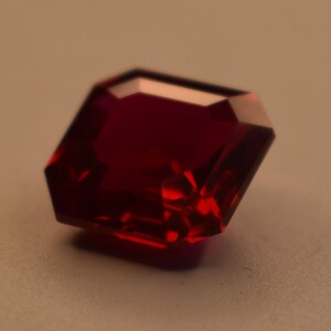 May include: A deep red, square-cut gemstone with faceted edges that catch the light. The stone features a translucent, saturated crimson hue and a polished surface, displaying a geometric shape with sharp corners and a clear, reflective quality.