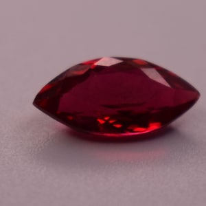 May include: A marquise-cut gemstone with a deep red colour and a faceted surface. The stone features a pointed oval shape with symmetrical edges, reflecting light across its polished, translucent, and vibrant crimson-toned crystalline structure.