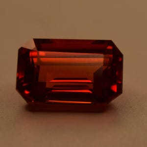 May include: A rectangular, emerald-cut gemstone with a deep, translucent red color. The stone features sharp, clean facets that create internal light reflections and shadows, showcasing a vibrant, saturated crimson hue throughout its structure.