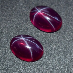May include: Two oval-shaped, deep red cabochon gemstones, each featuring a distinct six-rayed white star pattern across the smooth, polished surface. The stones are translucent and display a vibrant, saturated crimson hue with light reflecting off the curved tops.
