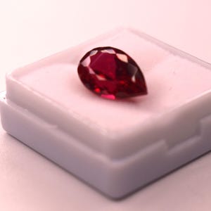 May include: A pear-shaped, deep red faceted gemstone resting inside a small, square white display box. The gemstone features multiple triangular facets that reflect light, creating a brilliant, polished appearance against the white interior.