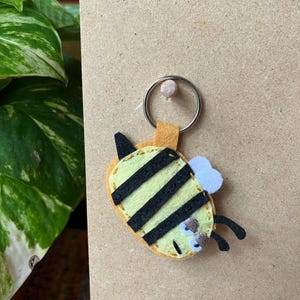 May include: A handmade felt bee keychain. The bee has a yellow body with black stripes, small black antennae, and a pair of white wings. The keychain includes a metal ring and a small tan felt tab.