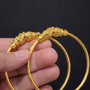 May include: Two gold-coloured bangles with a textured surface. Each bangle features a decorative head design at the top. The jewellery is displayed against a dark background, highlighting the bright gold colour.
