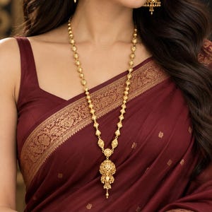May include: A gold necklace featuring a series of textured beads and a central bell-shaped pendant, paired with matching bell-shaped drop earrings. The jewelry set is worn with a deep maroon garment featuring a gold patterned border.
