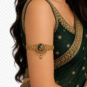 May include: A gold-toned armlet featuring an intricate filigree band with a central circular medallion. The medallion displays a dark green inlay with a gold floral motif, accented by small red stones and dangling white pearl-like beads.