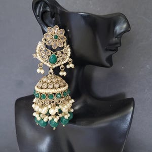May include: Gold-toned chandelier earrings with emerald green and clear accents. The earrings feature a floral design at the top, a teardrop-shaped emerald, and a bell-shaped bottom with dangling green beads and small white pearls. The earrings are displayed on a black mannequin head.