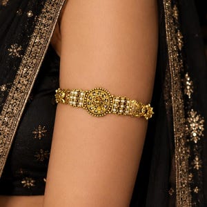 May include: A gold armlet featuring a central circular medallion with intricate filigree detailing, flanked by rows of small white pearls and gold beads. The metal has an antique finish, and the band is constructed with a series of connected segments.