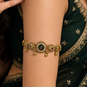 May include: An antique gold-toned armlet featuring a central circular medallion with a deep green enamel inlay and a small red stone. The band is decorated with intricate metalwork, small white beads, and dangling gold-toned charms along the edge.