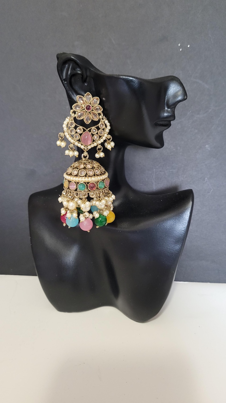 May include: An ornate gold-toned earring with a floral design and a pink gemstone. The earring features a bell-shaped bottom adorned with colorful beads in pink, green, blue, and yellow, and small white pearls.