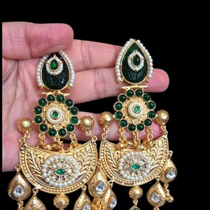 May include: A pair of gold-toned drop earrings with intricate detailing. The earrings feature green and white accents, including emerald-colored stones and small pearls. The design incorporates floral and teardrop shapes.