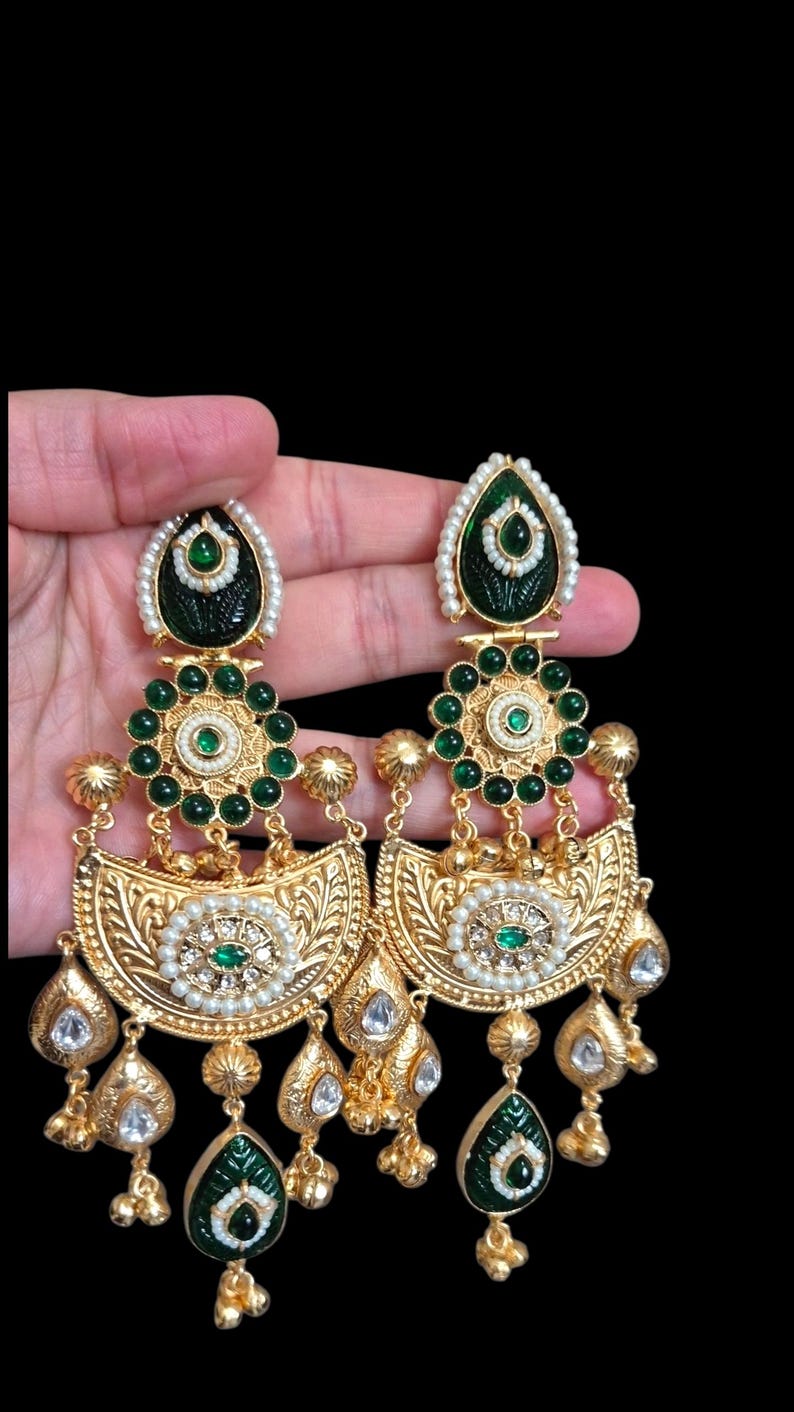 May include: A pair of gold-toned drop earrings with intricate detailing. The earrings feature green and white accents, including emerald-colored stones and small pearls. The design incorporates floral and teardrop shapes.