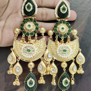 May include: Ornate gold-toned earrings with intricate detailing. Each earring features a teardrop-shaped green stone, surrounded by white accents and a gold frame. The design includes multiple layers of embellishments, including small green stones and pearl-like beads.