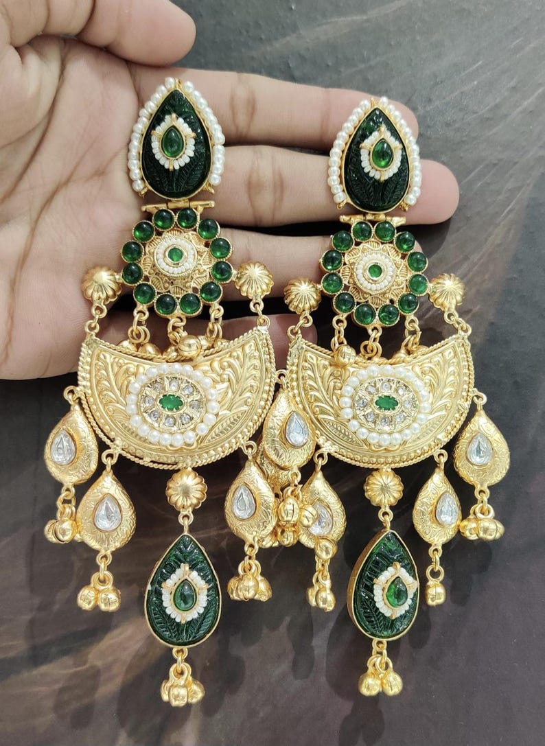 May include: Ornate gold-toned earrings with intricate detailing. Each earring features a teardrop-shaped green stone, surrounded by white accents and a gold frame. The design includes multiple layers of embellishments, including small green stones and pearl-like beads.