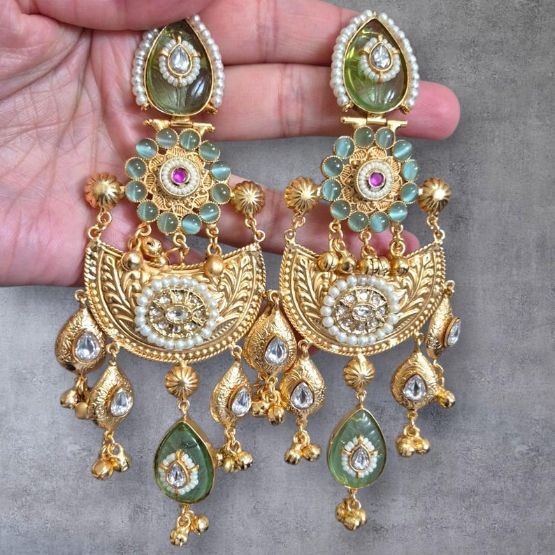 May include: A pair of ornate gold-colored earrings with intricate detailing. Each earring features a teardrop-shaped green stone, a central floral design with a pink center, and dangling accents. The earrings are embellished with pearls and clear stones.
