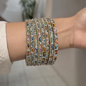 May include: A set of patterned bangles featuring floral designs and rhinestone embellishments. The bangles are predominantly white, adorned with a repeating pattern of red, blue, yellow, and green flowers. The bracelets are worn on the wrist.
