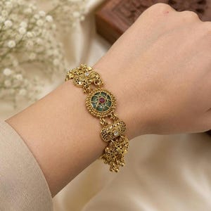 May include: A gold-toned metal bracelet featuring a central circular link with a green enamel inlay and a small red stone at its center. The bracelet includes ornate filigree rectangular links and dangling gold-toned charms along the chain.