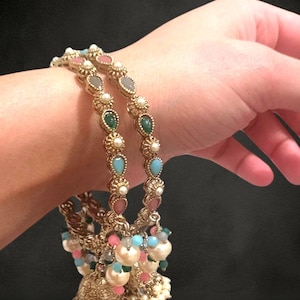 Puede incluir: Brazaletes dorados con piedras preciosas de colores y colgantes en forma de campana. Los brazaletes presentan detalles en rosa, azul, verde y perlas. Los colgantes están adornados con perlas y cuentas, creando un diseño elegante. Las joyas se exhiben sobre un fondo oscuro.