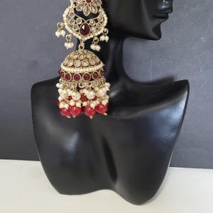 May include: An ornate gold-toned earring with a floral design and red and clear gemstones. The earring features a bell-shaped bottom with red beads and white pearls. The earring is displayed on a black mannequin head.