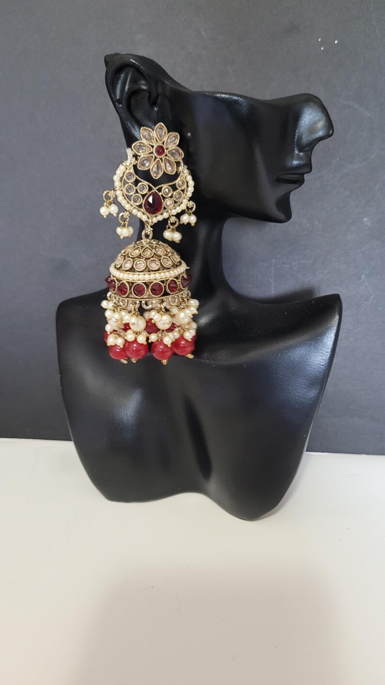 May include: An ornate gold-toned earring with a floral design and red and clear gemstones. The earring features a bell-shaped bottom with red beads and white pearls. The earring is displayed on a black mannequin head.