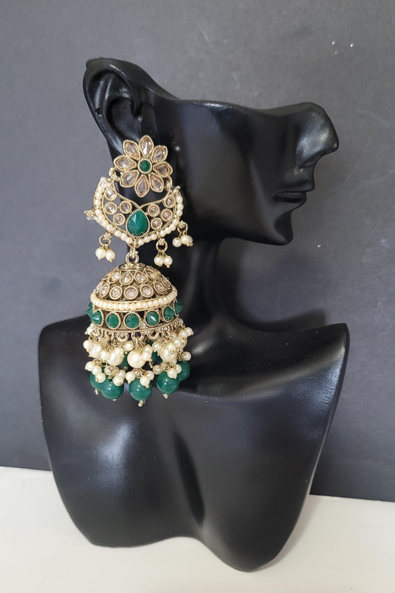 May include: Gold-toned chandelier earrings with emerald green and clear accents. The earrings feature a floral design at the top, a teardrop-shaped emerald, and a bell-shaped bottom with dangling green beads and small white pearls. The earrings are displayed on a black mannequin head.