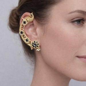 May include: A gold ear cuff with a peacock motif. The cuff curves around the ear, embellished with black and blue gemstones and small pearls. The peacock's head is at the top, with a circular design at the bottom.