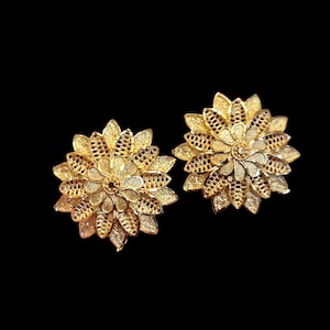 May include: A pair of gold-colored flower-shaped earrings. Each earring features intricate petal detailing and a textured center, set against a black background. The earrings are designed to resemble a blooming flower.