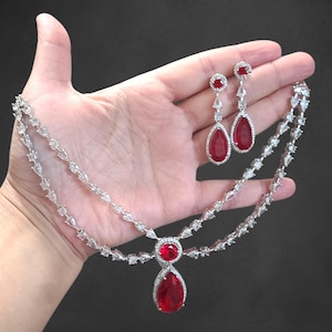 Può includere: Un set di collana e orecchini in argento con grandi pietre preziose rosse a forma di goccia. La collana ha un design a doppia fila con pietre chiare più piccole. Gli orecchini sono lunghi e pendenti.