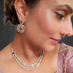 May include: A two-strand necklace and matching earrings set. The jewellery features a square, ruby-coloured centre stone surrounded by a halo of smaller, clear stones. The earrings are square with a floral design. The necklace has a similar design.