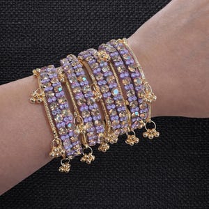May include: A collection of gold-toned bangles adorned with lavender and champagne-colored rhinestones. Each bangle features a row of sparkling stones and dangling gold charms, creating a glamorous look. The jewelry is displayed on a dark background.