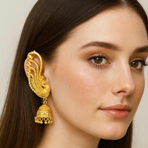 Peut inclure: Un ear cuff doré orné d'un motif de paon. L'ear cuff s'enroule autour de l'oreille et comporte un charm en forme de cloche suspendue avec de petits accents ronds. Le bijou est orné et détaillé.