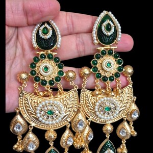May include: Gold-toned chandelier earrings with intricate detailing. The earrings feature emerald green accents, small white pearls, and clear, teardrop-shaped gems. The design includes floral and leaf motifs, creating a luxurious and ornate appearance.