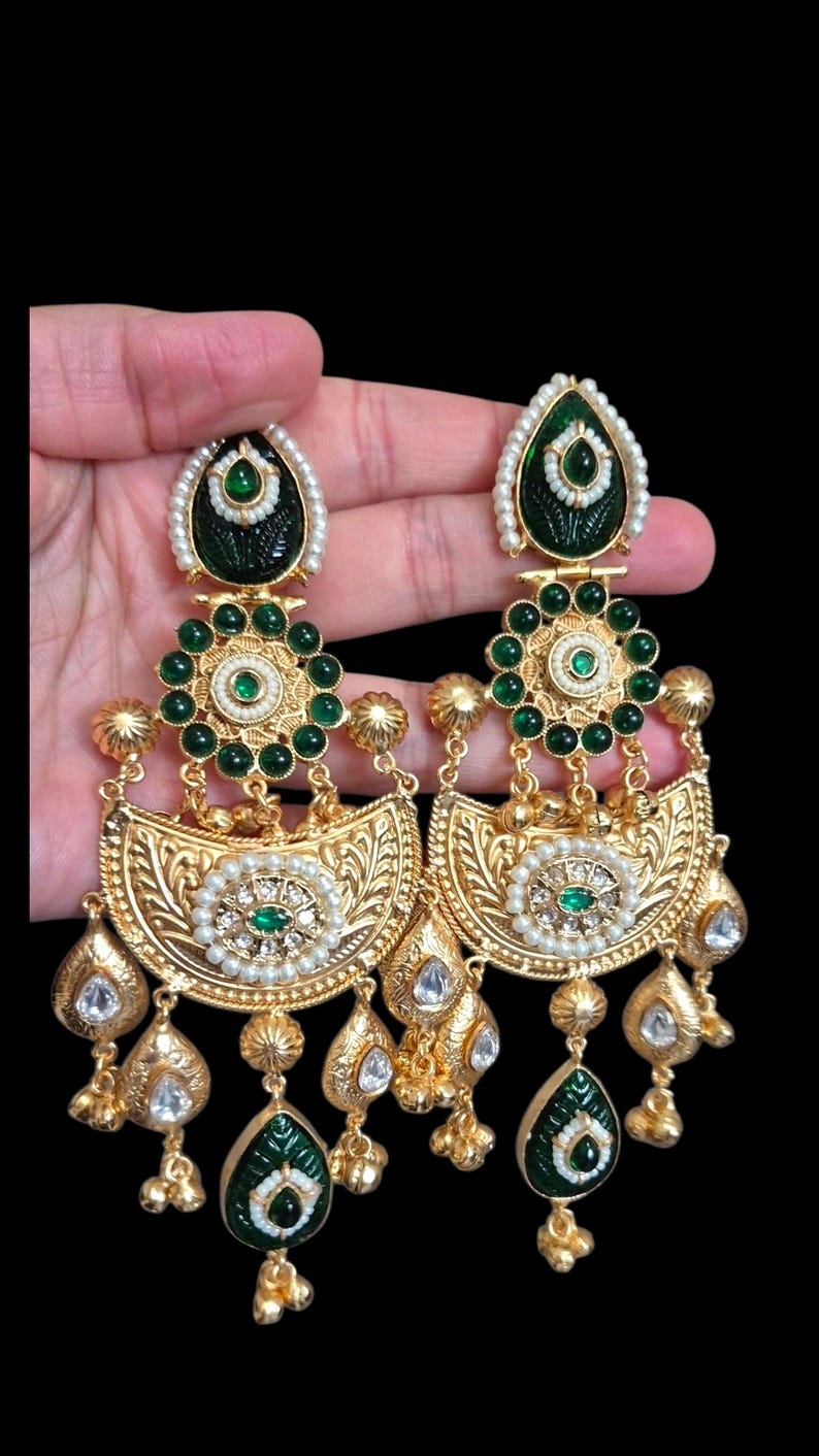 May include: Gold-toned chandelier earrings with intricate detailing. The earrings feature emerald green accents, small white pearls, and clear, teardrop-shaped gems. The design includes floral and leaf motifs, creating a luxurious and ornate appearance.