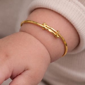 Op de afbeelding: Een gouden babyarmband met een getextureerd oppervlak. De armband is verstelbaar en heeft een sluiting. De armband wordt getoond op de pols van een baby en de achtergrond is een zachte, neutrale kleur.