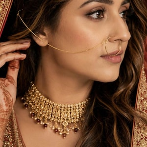 May include: A gold-toned choker necklace featuring intricate metalwork, small white pearls, and deep red beads. A matching gold nose ring is connected by a thin chain to a gold headpiece adorned with white pearls and red beads.
