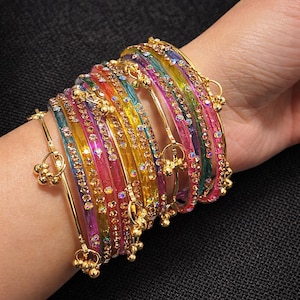 May include: A selection of vibrant, multi-coloured bangles, including pink, yellow, green, and blue, embellished with tiny, glittering gemstones. The bangles are stacked on a wrist, with gold-coloured details and hanging charms.