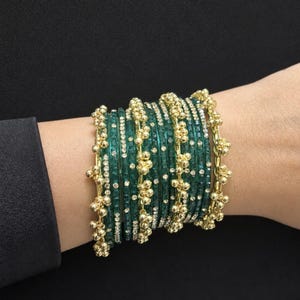 Könnte beinhalten: Ein Set grüner Armreifen mit goldenen Akzenten und eingebetteten Strasssteinen. Die Armreifen sind auf einem Handgelenk gestapelt und zeigen ihr aufwendiges Design. Die goldenen Akzente weisen kleine, kugelförmige Verzierungen auf, die dem Schmuck einen Hauch von Eleganz verleihen.