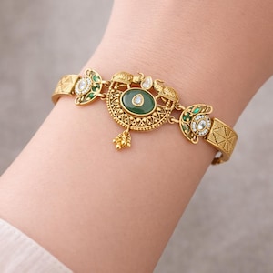 Peut inclure: Bracelet doré orné d'un motif élaboré avec des accents verts et blancs. L'élément central présente une grande pierre verte ovale, entourée de détails complexes et de petites décorations pendantes. Le bracelet est porté au poignet.