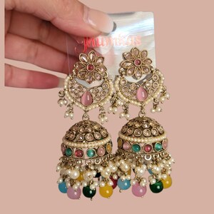 May include: A pair of gold-toned jhumka earrings with intricate detailing. The earrings feature a floral design at the top, a teardrop-shaped pink stone, and a bell-shaped bottom adorned with colorful beads and pearls. The text "Mumkas" is visible.