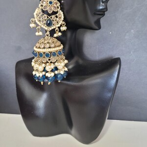 May include: Gold-toned, ornate earrings with blue and white accents. The earrings feature a floral design at the top, a bell-shaped body, and dangling blue beads. The earrings are displayed on a black mannequin head.