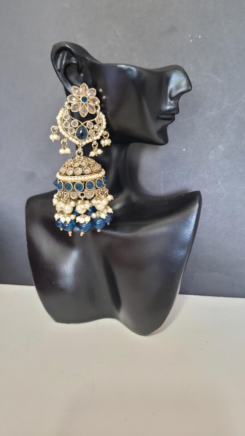 May include: Gold-toned, ornate earrings with blue and white accents. The earrings feature a floral design at the top, a bell-shaped body, and dangling blue beads. The earrings are displayed on a black mannequin head.