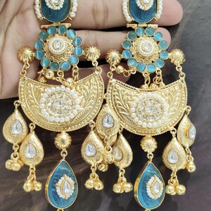 May include: Ornate gold-toned earrings with turquoise and white accents. The earrings feature a teardrop-shaped turquoise stone at the bottom, a floral design, and a pearl border. The earrings are held up by a hand.