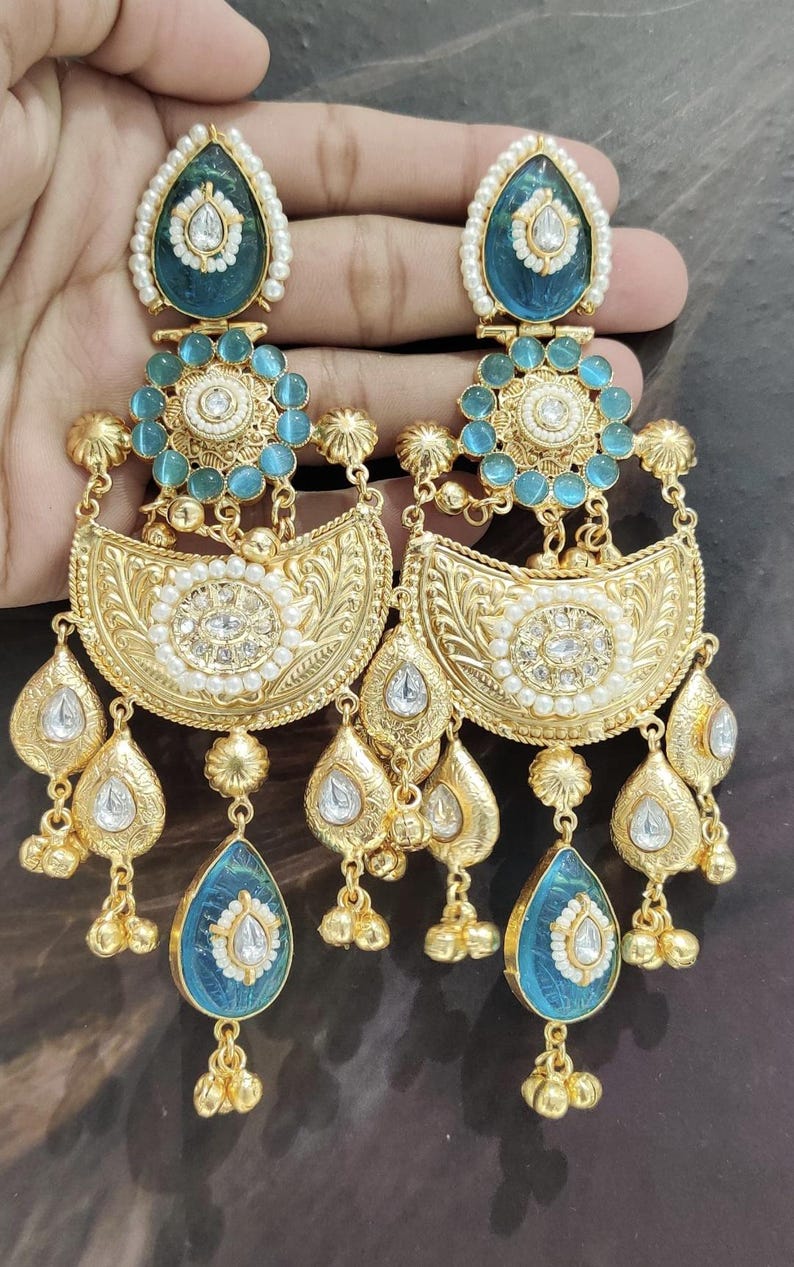 May include: Ornate gold-toned earrings with turquoise and white accents. The earrings feature a teardrop-shaped turquoise stone at the bottom, a floral design, and a pearl border. The earrings are held up by a hand.