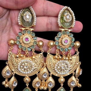 May include: Gold-toned chandelier earrings with intricate detailing. Each earring features a pear-shaped green stone, surrounded by pearls and clear crystals. The design includes floral and teardrop elements, with a central cluster of green stones and a pearl-lined top.