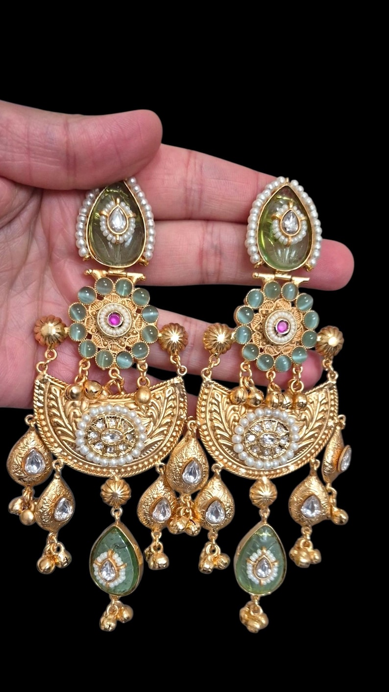 May include: Gold-toned chandelier earrings with intricate detailing. Each earring features a pear-shaped green stone, surrounded by pearls and clear crystals. The design includes floral and teardrop elements, with a central cluster of green stones and a pearl-lined top.